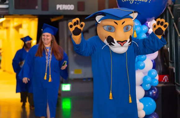 A UMA graduate celebrates at a commencement ceremony with UMA mascot Journey the Puma