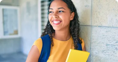 a student wearing a backpack and holding a book while smiling and leaning against a wall