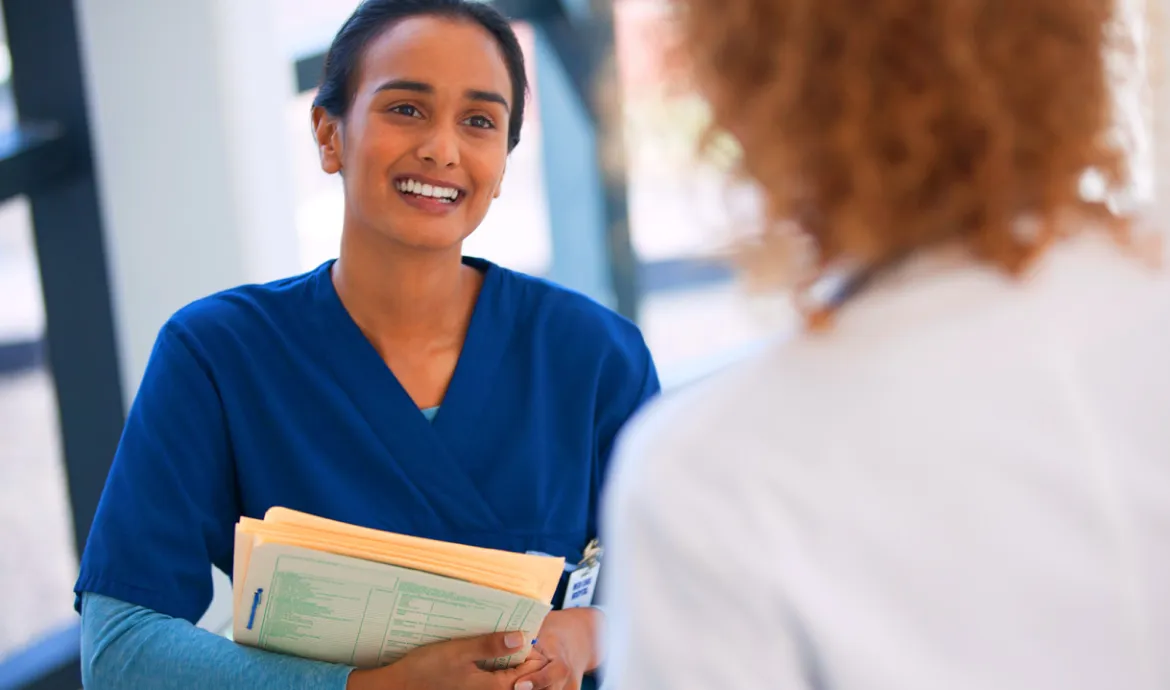 a medical assistant smiling while holding patient files and talking to a physician