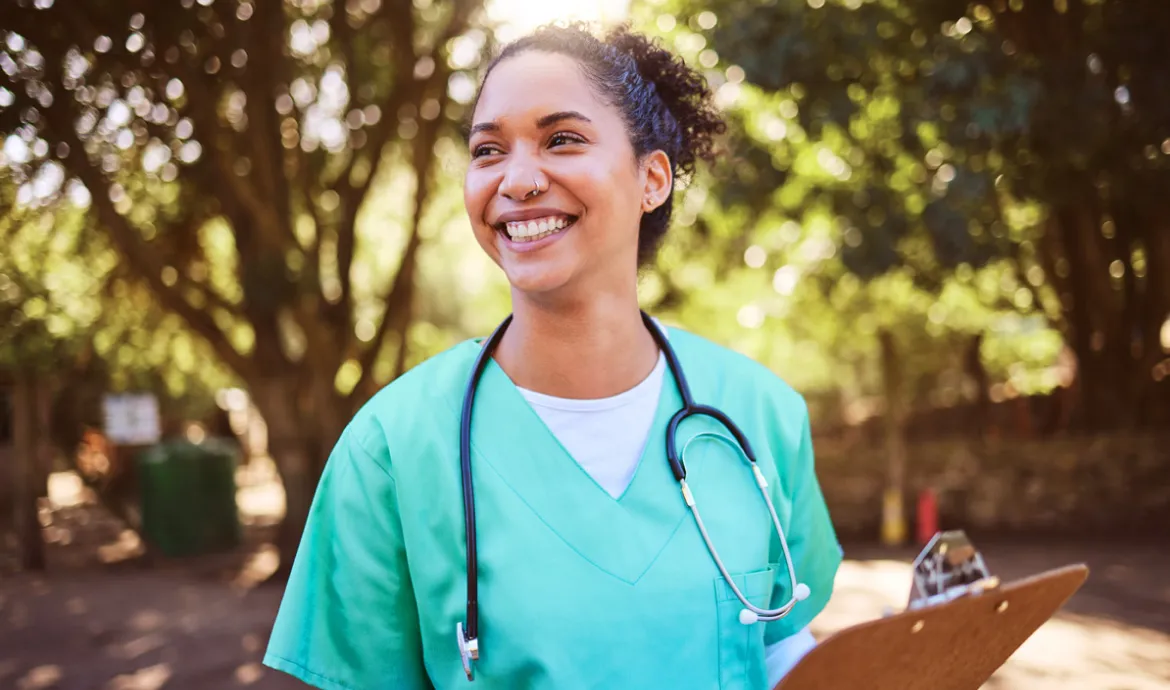 a nursing assistant smiling while holding a clipboard and standing in a park