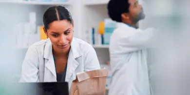 pharmacy technician working on a computer while another looks at drugs on a shelf