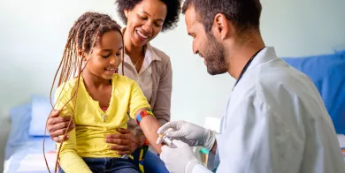 phlebotomy technician holding vial while smiling at a child who is sitting on her mom’s lap