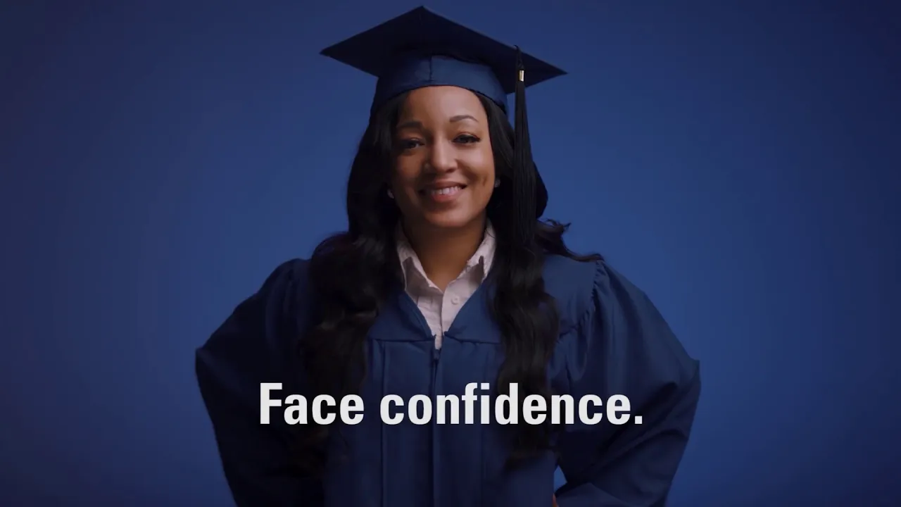 A UMA graduate smiles while wearing cap and gown. This is a poster image that displays before showing an embedded YouTube video.